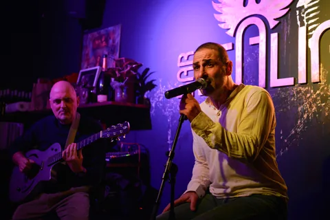 Sänger am Mikrofon vor der beleuchteten Bar-Italia-Logowand, Band im Halbdunkel
