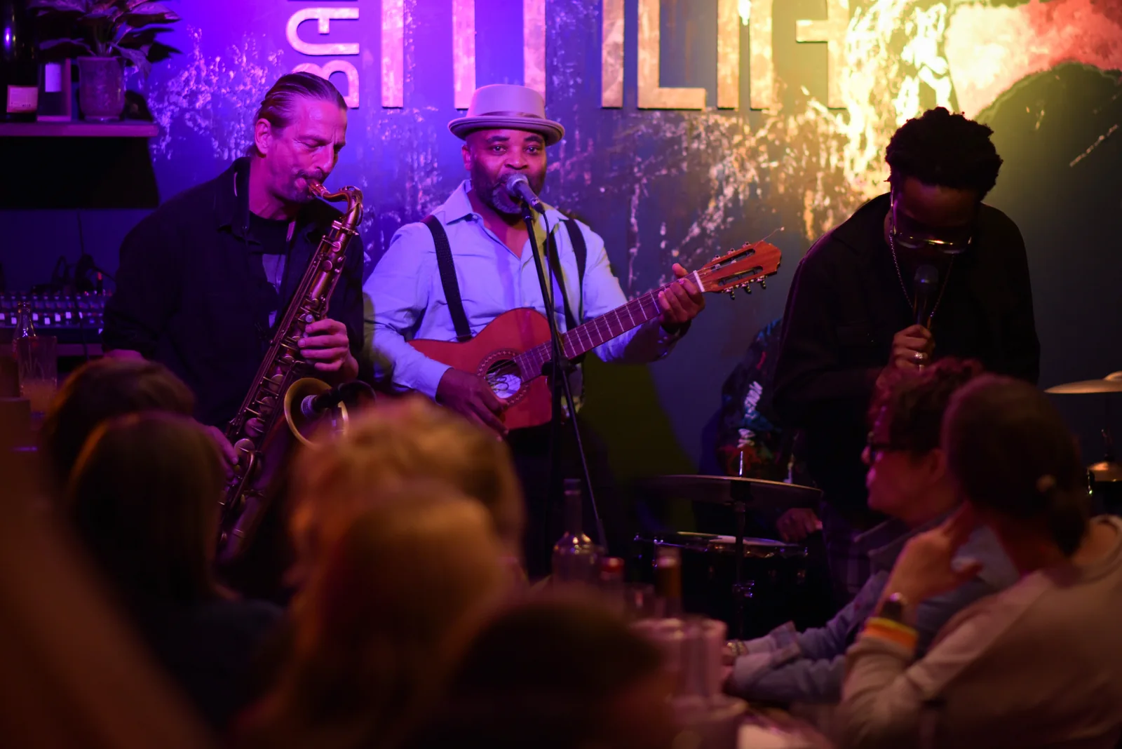 Trio mit Saxofon, Gitarre und Gesang auf der Bühne, Gäste an den Tischen davor