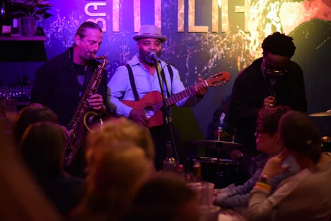 Trio mit Saxofon, Gitarre und Gesang auf der Bühne, Gäste an den Tischen davor